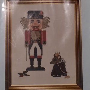 May include: A cross-stitch pattern titled "Nutcracker" by Kingsland. The design features a framed image of a nutcracker soldier and a mouse. The pattern is in a plastic sleeve.