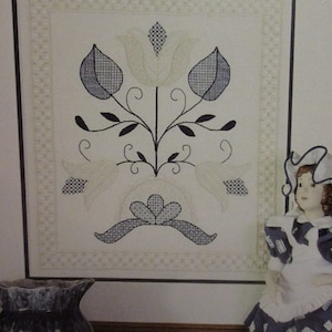 May include: A framed floral design featuring a central flower with blue and white patterned leaves and a matching bow. The design is framed with a decorative border. A doll in a blue and white dress is to the right.