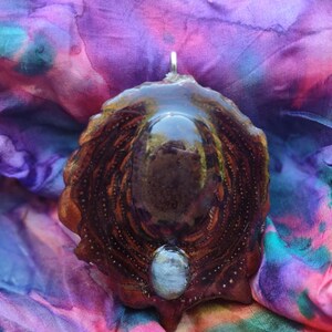 Puede incluir: Un colgante artesanal con un diseño orgánico único. El colgante presenta una base texturizada de color marrón oscuro con un centro brillante y oscuro y una pequeña piedra ovalada de color claro en la parte inferior. El colgante cuelga de una fianza plateada.