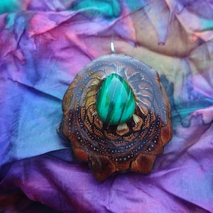 Puede incluir: Colgante artesanal con una piedra de malaquita verde engastada en una base de madera tallada. El colgante tiene un diseño detallado y texturizado y una argolla plateada. El fondo es una tela teñida con efecto tie-dye.
