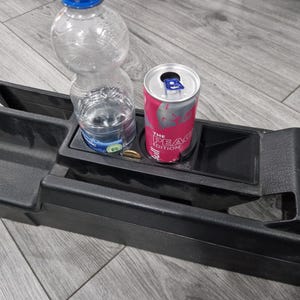 Könnte beinhalten: Schwarze Autokonsole mit eingebauten Getränkehaltern und Ablagefächern. Eine transparente Plastikwasserflasche und eine rosa Dose des Getränks The Peach Edition befinden sich in den Getränkehaltern. Eine Münze ist ebenfalls sichtbar.