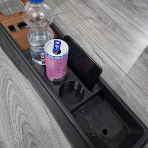 Puede incluir: Consola de coche negra con portavasos incorporado, ranura para teléfono y compartimento de almacenamiento. Una botella de agua de plástico transparente y una lata de bebida energética rosa están en la consola. Un teléfono inteligente negro está en la ranura para teléfono.