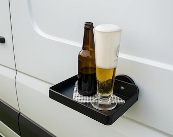 Puede incluir: Una bandeja negra montada en la pared con una botella de cerveza marrón y un vaso de cerveza con espuma. La bandeja está fijada a una superficie blanca y tiene una pequeña servilleta a cuadros.