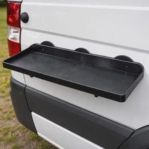 Könnte beinhalten: Ein schwarzes, klappbares Tablett, das an der Rückseite eines weißen Transporters befestigt ist. Das Tablett ist rechteckig mit abgerundeten Ecken und wird mit Saugnäpfen befestigt. Das Rücklicht des Transporters und ein Teil der Stoßstange sind sichtbar.