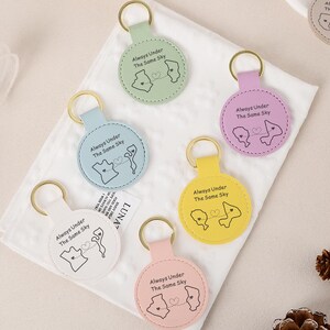 Puede incluir: Una colección de llaveros coloridos con anillos dorados. Cada llavero presenta un diseño circular con un gráfico y el texto "Always Under The Same Sky". Los llaveros están disponibles en varios colores, incluyendo marrón, negro, rojo y tonos pastel.