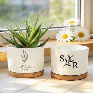 Puede incluir: Dos macetas de cerámica blanca con platillos de bambú. Una maceta tiene las iniciales "k + h" y la fecha "09.23.2025", la otra "S R". Ambas tienen un diseño floral negro y contienen plantas verdes. Las macetas están sobre una superficie blanca.