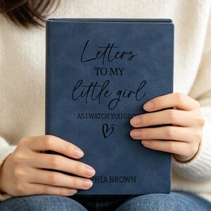 Może przedstawiać: Granatowy dziennik o matowym wykończeniu z wygrawerowanym czarnym napisem "Letters TO MY little girl AS I WATCH YOU GROW", małą ikoną serca oraz imieniem "SOPHIA BROWN" nadrukowanym w dolnej części przedniej okładki.