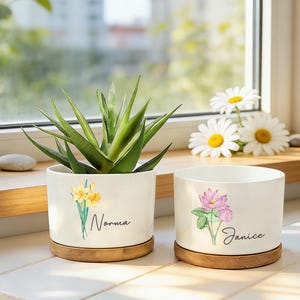 May include: Two white cylindrical ceramic planters with bamboo saucers. The left planter features a yellow daffodil illustration and the name Norma. The right planter features a pink lotus flower illustration and the name Janice. A green succulent sits in the left planter.