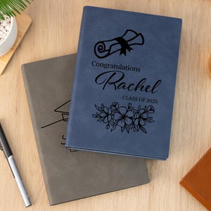 May include: Two graduation journals, one blue and one grey, with personalised text. The blue journal has the text "Congratulations Rachel Class of 2025" and a graduation cap and flowers design. The grey journal has a graduation cap design.