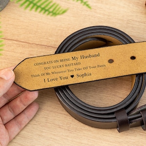 以下が含まれることがあります： 先端にパーソナライズされたメッセージが刻まれた茶色の革ベルト。テキストは「CONGRATS ON BEING My Husband YOU LUCKY BASTARD Think Of Me Whenever You Take Off Your Pants I Love You Sophia」と書かれています。ベルトは丸まっています。