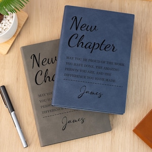 Può includere: Due diari rilegati in pelle, uno blu e uno grigio, con le parole "New Chapter" e un messaggio personalizzato. Una penna nera è appoggiata accanto ai diari su una superficie di legno. Sono visibili anche una piccola pianta e un bicchiere di liquido.