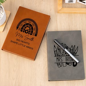 Puede incluir: Dos cuadernos personalizados. Uno es marrón con un diseño de arcoíris, el nombre "Mrs. Smith" y el texto "Big Hearts Shape Little Minds". El otro es gris con un bolígrafo y el nombre "Mrs. Brown".