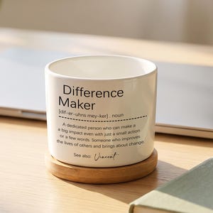 May include: A white ceramic mug with the text "Difference Maker" in black, on a wooden coaster. The mug features a definition. A laptop and a green notebook are visible in the background.