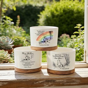 Può includere: Tre vasi di ceramica bianca con basi in legno, ognuno con una diversa illustrazione e citazione di Winnie the Pooh. I vasi sono cilindrici e poggiano su una superficie di legno.