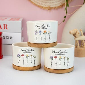 May include: Three white ceramic Mom's Garden planters with bamboo saucers. Each planter features a floral design with names like "sherry" and "julie" below. The text "Mom's Garden LOVE GROWS HERE" is printed on each planter.