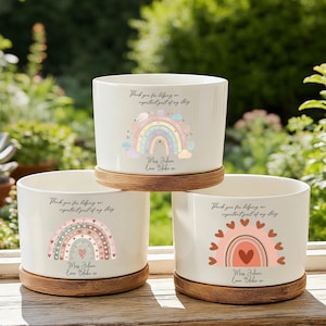 Può includere: Tre vasi cilindrici in ceramica bianca, ciascuno poggiato su una base circolare in legno. Ogni vaso presenta una grafica arcobaleno unica con cuori e il testo stampato: Thank you for helping me grow, important part of my story, Miss Juhan, Love Blake xx.