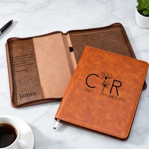 Op de afbeelding: Een bruine lederen portfolio met ritssluiting, voorzien van de zwart gedrukte naam CINDY RASMUSSEN en een bloemmotief tussen de letters C en R. De open binnenzijde toont een donkerbruin vak met de naam James en een gedrukte tekst.