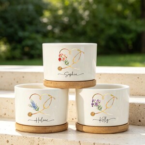Puede incluir: Tres macetas cilíndricas de cerámica blanca, cada una sobre una base circular de bambú. Cada maceta presenta un gráfico de estetoscopio dorado con forma de corazón, decorado con ilustraciones florales y los nombres Sophia, Helene y Kelly.