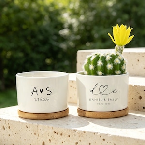 Pode incluir: Dois vasos cilíndricos de cerâmica branca com bases de bambu. O vaso esquerdo apresenta o texto A coração S e 1.15.25. O vaso direito contém um cacto verde com uma flor amarela e o texto d coração e, DANIEL & EMILY, 06.12.2025.