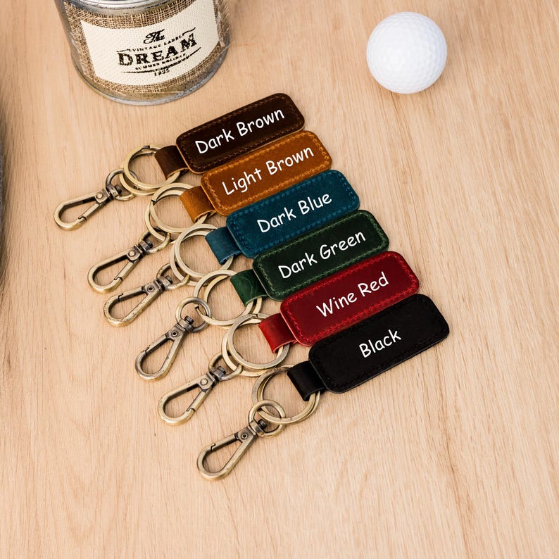 May include: A selection of leather keyrings in shades of dark brown, light brown, dark blue, dark green, wine red, and black. Each keyring features a metal clasp and a tag with the colour name in white text. A golf ball and a jar are in the background.