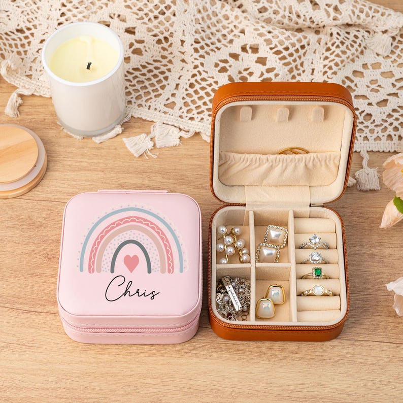 May include: A pink jewelry box with a rainbow and heart design. The box has the name "Chris" printed on it. The box is open and shows a brown interior with compartments for rings, earrings, and other jewelry.