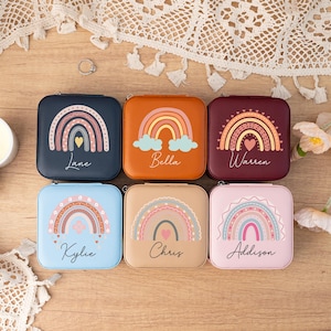 May include: Six square-shaped jewelry boxes with rainbow designs and personalized names. The boxes are in various colors, including blue, brown, burgundy, and pink. Each box features a rainbow with a heart in the center and a name written below.