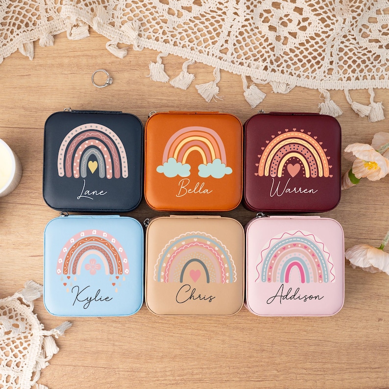 May include: Six square-shaped jewelry boxes with rainbow designs and personalized names. The boxes are in various colors, including blue, brown, burgundy, and pink. Each box features a rainbow with a heart in the center and a name written below.