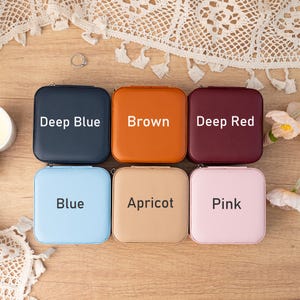 May include: Six square-shaped jewelry boxes with different colors. The boxes are arranged in two rows of three. The top row has boxes labeled "Deep Blue", "Brown", and "Deep Red". The bottom row has boxes labeled "Blue", "Apricot", and "Pink".