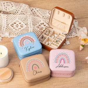 May include: Three small, square, personalized jewelry boxes with rainbow designs. The boxes are blue, pink, and brown. Each box has a name on it: Kylie, Addison, and Chris.