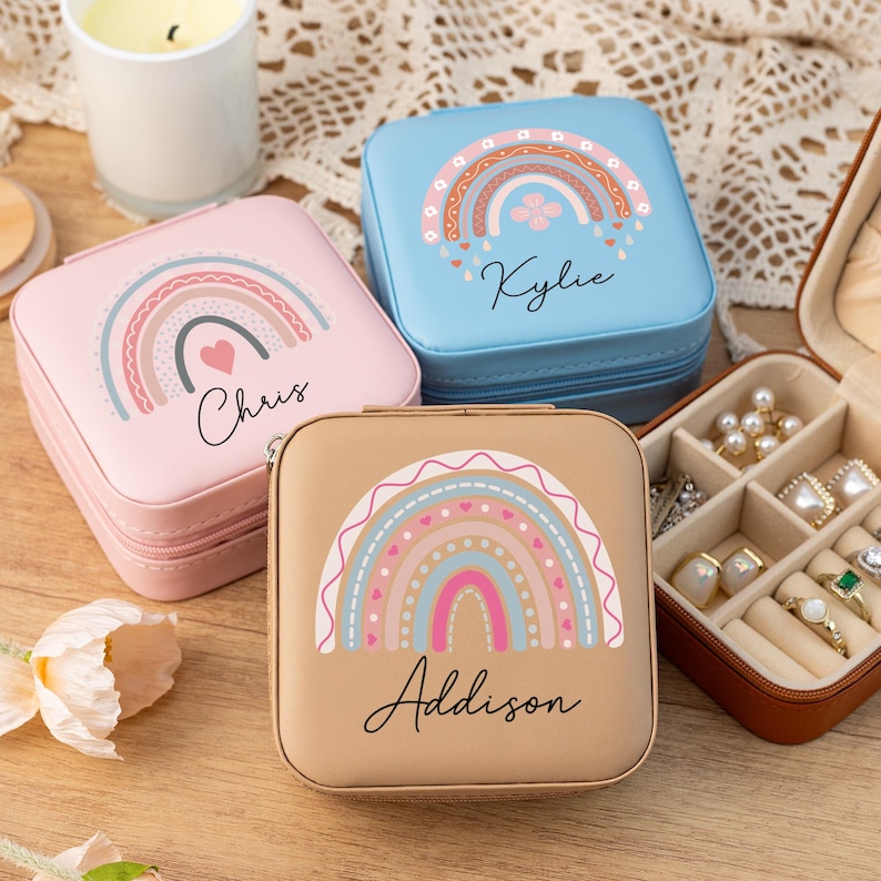 May include: Three small square jewelry boxes with rainbow designs and personalized names. The boxes are blue, pink, and brown. The boxes are on a wooden surface.
