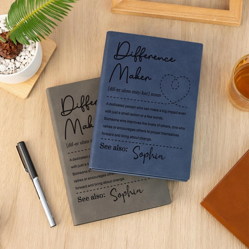 Pode incluir: Dois cadernos, um azul e outro cinzento, com as palavras "Difference Maker" e uma defini&ccedil;&atilde;o impressa na capa. O caderno azul tem um desenho em forma de cora&ccedil;&atilde;o. Uma caneta preta repousa ao lado dos cadernos.