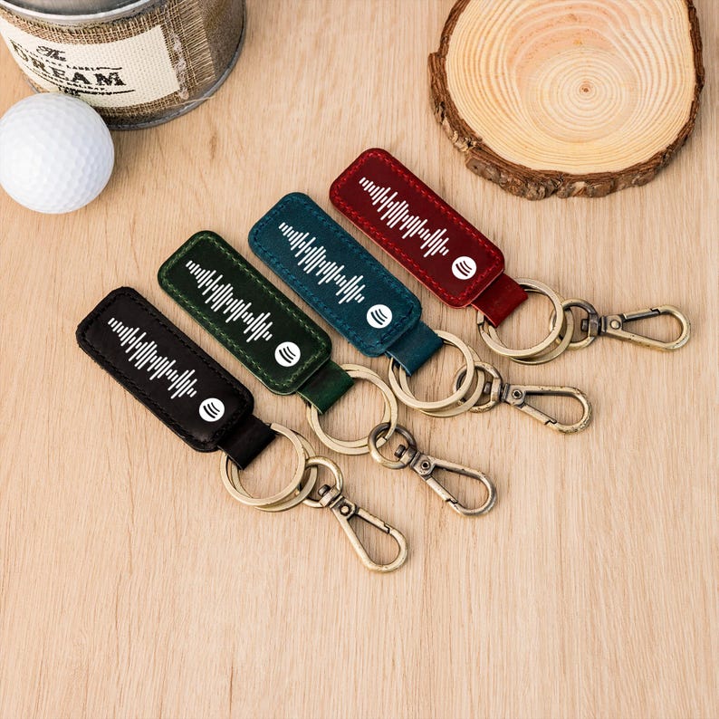 May include: Four leather keyrings in black, green, teal, and red. Each keyring features a white sound wave design and a white circular logo. The keyrings have gold-coloured metal rings and clasps, and are arranged on a wooden surface.