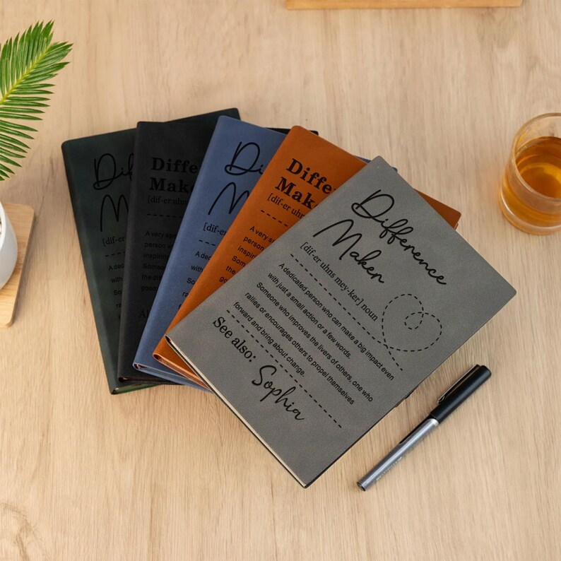 Pode incluir: Uma cole&ccedil;&atilde;o de cadernos em v&aacute;rias cores, incluindo cinza, preto, azul e laranja. Cada caderno apresenta o texto "Difference Maker" e uma defini&ccedil;&atilde;o. Uma caneta preta est&aacute; ao lado dos cadernos.