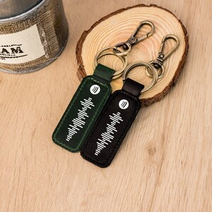 May include: Two keyrings with a dark green and black leather tag. Each tag features a white sound wave design and a metal clasp. The keyrings are on a wooden surface.