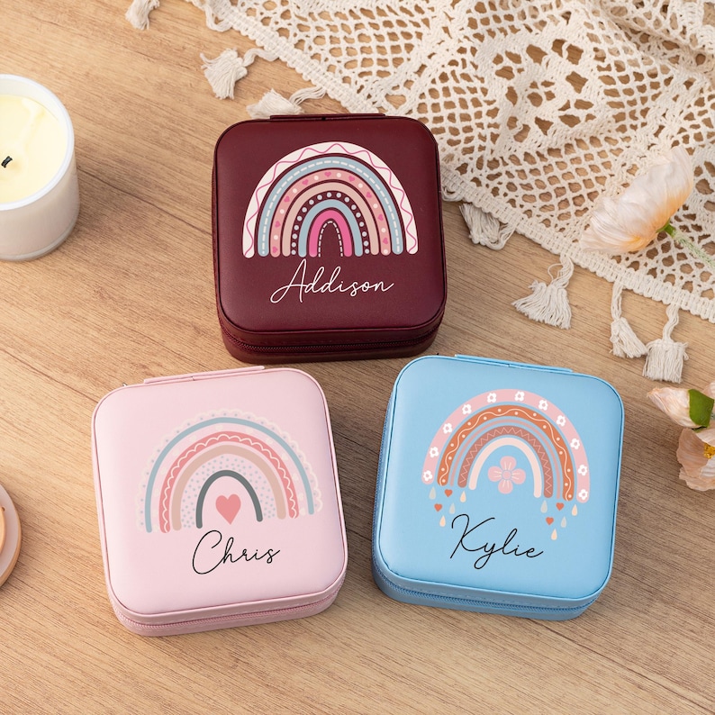 May include: Three small square jewelry boxes with rainbow designs. The boxes are burgundy, pink, and blue. Each box has a name on it: Addison, Chris, and Kylie.