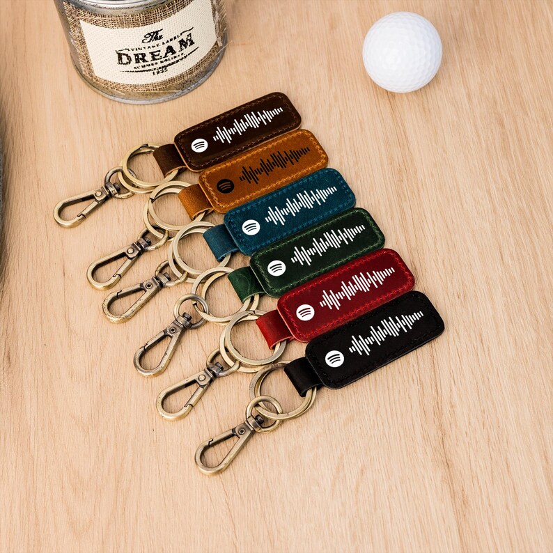 May include: A selection of leather keyrings in different colours, each with a white sound wave design and a Spotify logo. The keyrings have metal clasps and rings. A golf ball and a jar are in the background.