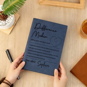 Pode incluir: Um caderno azul com as palavras "Difference Maker" e uma defini&ccedil;&atilde;o impressa na capa. O caderno tamb&eacute;m inclui o nome "Sophia". Uma caneta e uma pequena planta s&atilde;o vis&iacute;veis no fundo.