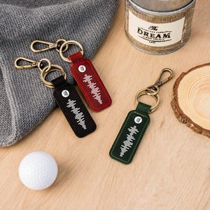 May include: Leather keyrings in black, red, and green with gold-coloured clasps. Each keyring displays a white sound wave design and a circular logo. A white golf ball and a grey knitted fabric are also in the image.
