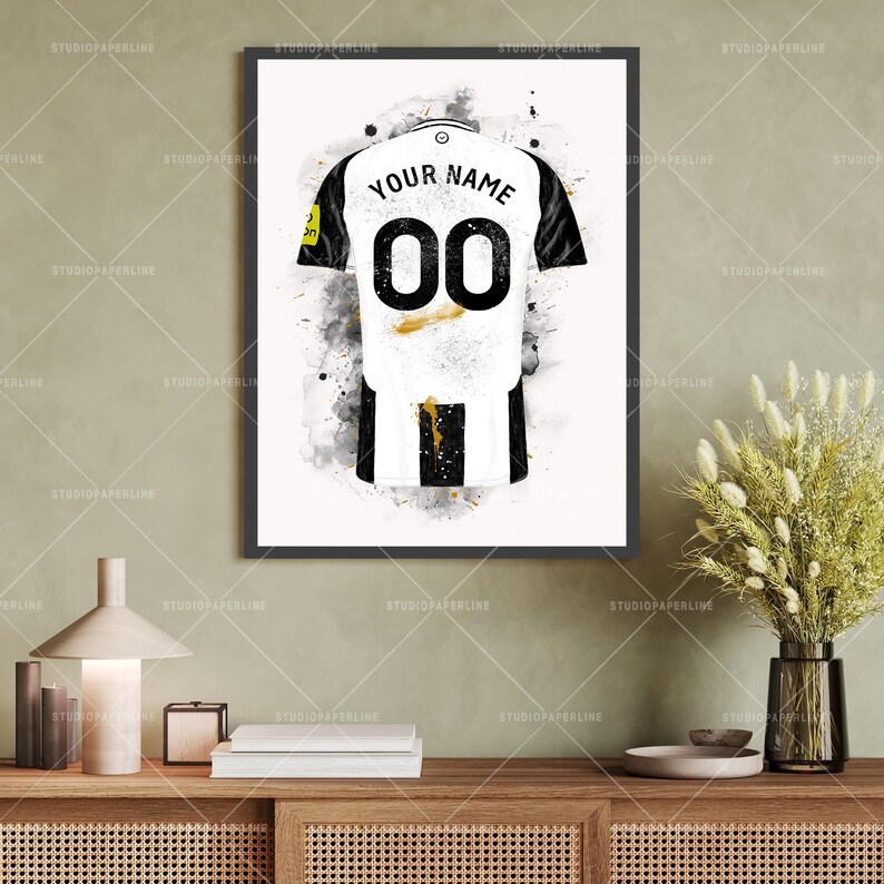 May include: Framed art print of a soccer jersey with the text "YOUR NAME" above the number "00". The jersey is white with black accents and a black and white striped bottom. Gold accents and paint splatters are visible.