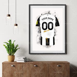 May include: A framed art print of a soccer jersey, featuring a white and black design with the number "00" on the back. The words "YOUR NAME" are above the number. The artwork has a watercolor effect with gold accents.