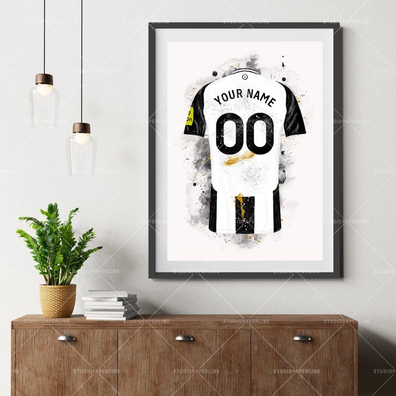 May include: A framed art print of a soccer jersey, featuring a white and black design with the number "00" on the back. The words "YOUR NAME" are above the number. The artwork has a watercolor effect with gold accents.
