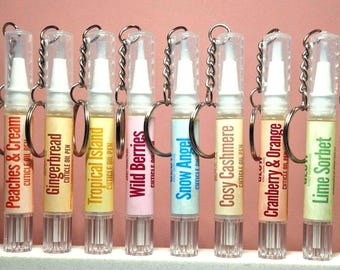 May include: A row of eight clear plastic cuticle oil pens with silver keychains. Each pen features a label with a unique scent name: Peaches & Cream, Gingerbread, Tropical Island, Wild Berries, Snow Angel, Cosy Cashmere, Cranberry & Orange, and Lime Sorbet.