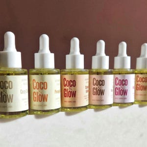 Puede incluir: Una fila de frascos cuentagotas de vidrio llenos de un líquido amarillo. Cada frasco tiene una tapa blanca y una etiqueta con las palabras "Coco & Glow" en una fuente estilizada. Las etiquetas son de varios colores, incluyendo beige, naranja y rosa. Los frascos son para el cuidado de la piel.