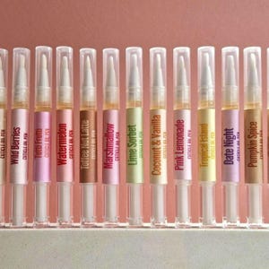 Puede incluir: Una fila de doce bolígrafos de aceite para cutículas de plástico transparente con puntas de pincel blancas. Cada bolígrafo tiene una etiqueta de color con texto que incluye Wild Berries, Tutti Frutti, Watermelon, Toffee Nut Latte, Marshmallow, Lime Sorbet, Coconut & Vanilla, Pink Lemonade, Tropical Island, Date Night y Pumpkin Spice.