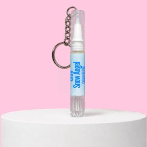 Puede incluir: Un bolígrafo de aceite para cutículas de plástico transparente con un llavero de metal plateado unido a la tapa. La etiqueta es azul claro con texto azul oscuro que dice Snow Angel Cuticle Oil Pen. La punta aplicadora es blanca.