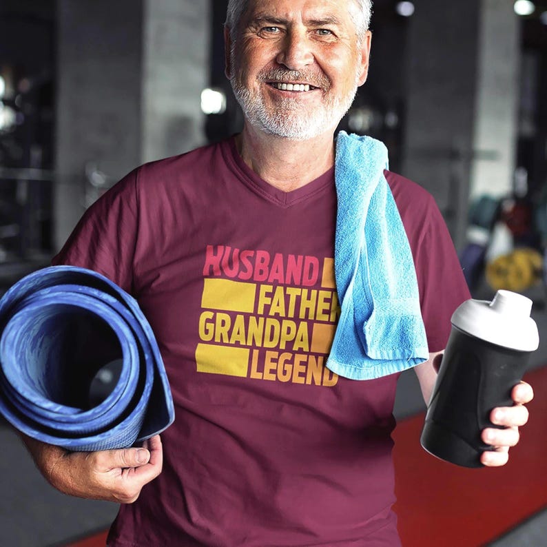 May include: A maroon V-neck t-shirt with the words "HUSBAND FATHER GRANDPA LEGEND" in a yellow and pink font. The person is holding a blue yoga mat, a blue towel, and a black and white water bottle.