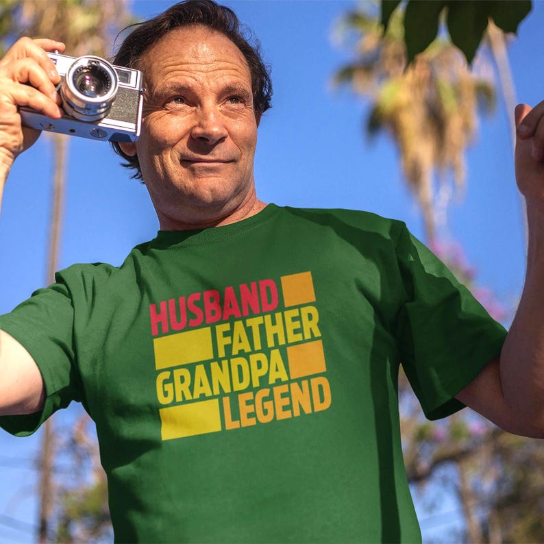 May include: Green t-shirt with the words "HUSBAND FATHER GRANDPA LEGEND" in a stacked design. The text is in red, orange, and yellow. The person in the image is holding a vintage camera.