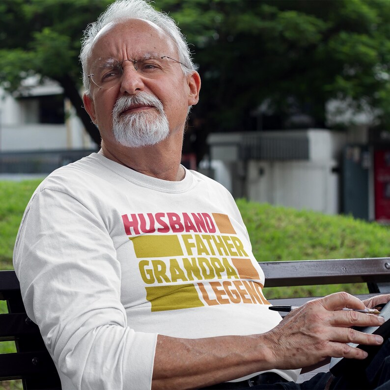May include: White long-sleeve shirt with the words "HUSBAND FATHER GRANDPA LEGEND" in a layered, rectangular design. The text is in red, orange, and yellow. The shirt is worn by a person with a white beard and glasses.