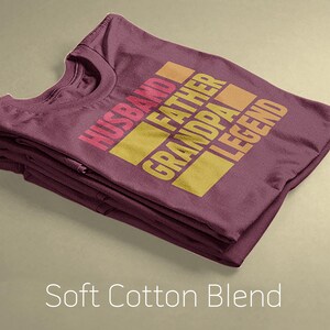 May include: Folded maroon t-shirt with the words "HUSBAND FATHER GRANDPA LEGEND" in a gradient of pink, orange, and yellow. The words are stacked in a rectangular design. The text "Soft Cotton Blend" is at the bottom.