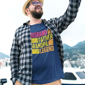 May include: Navy blue t-shirt with the words "HUSBAND FATHER GRANDPA LEGEND" in a colorful, block-letter design. The person in the image is wearing a plaid shirt and a hat.